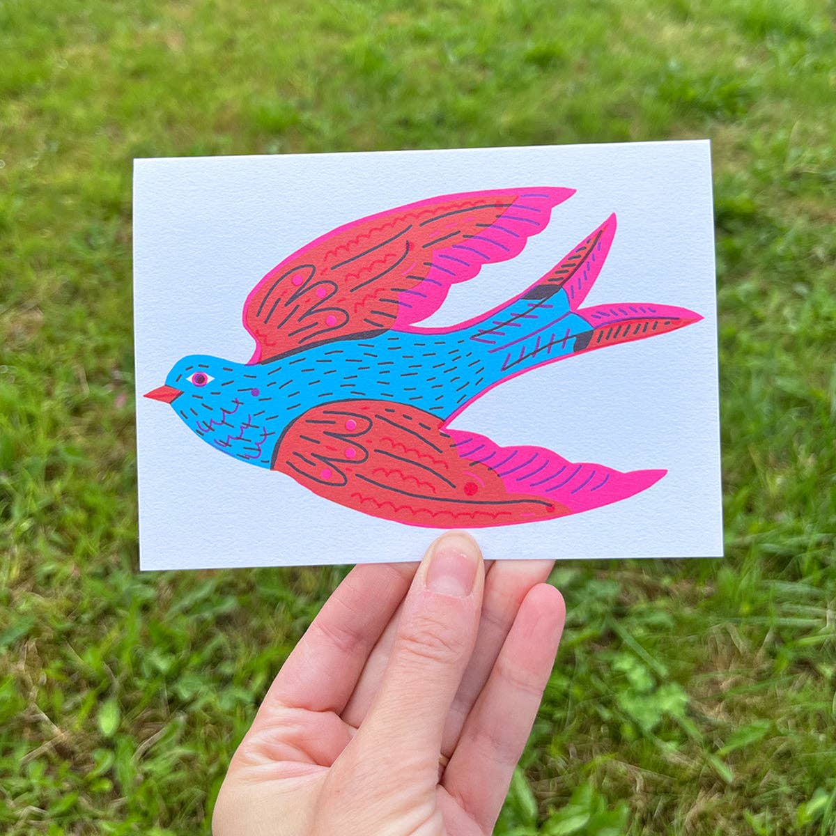 Hand holding a card with a colorful bird design against a grassy background