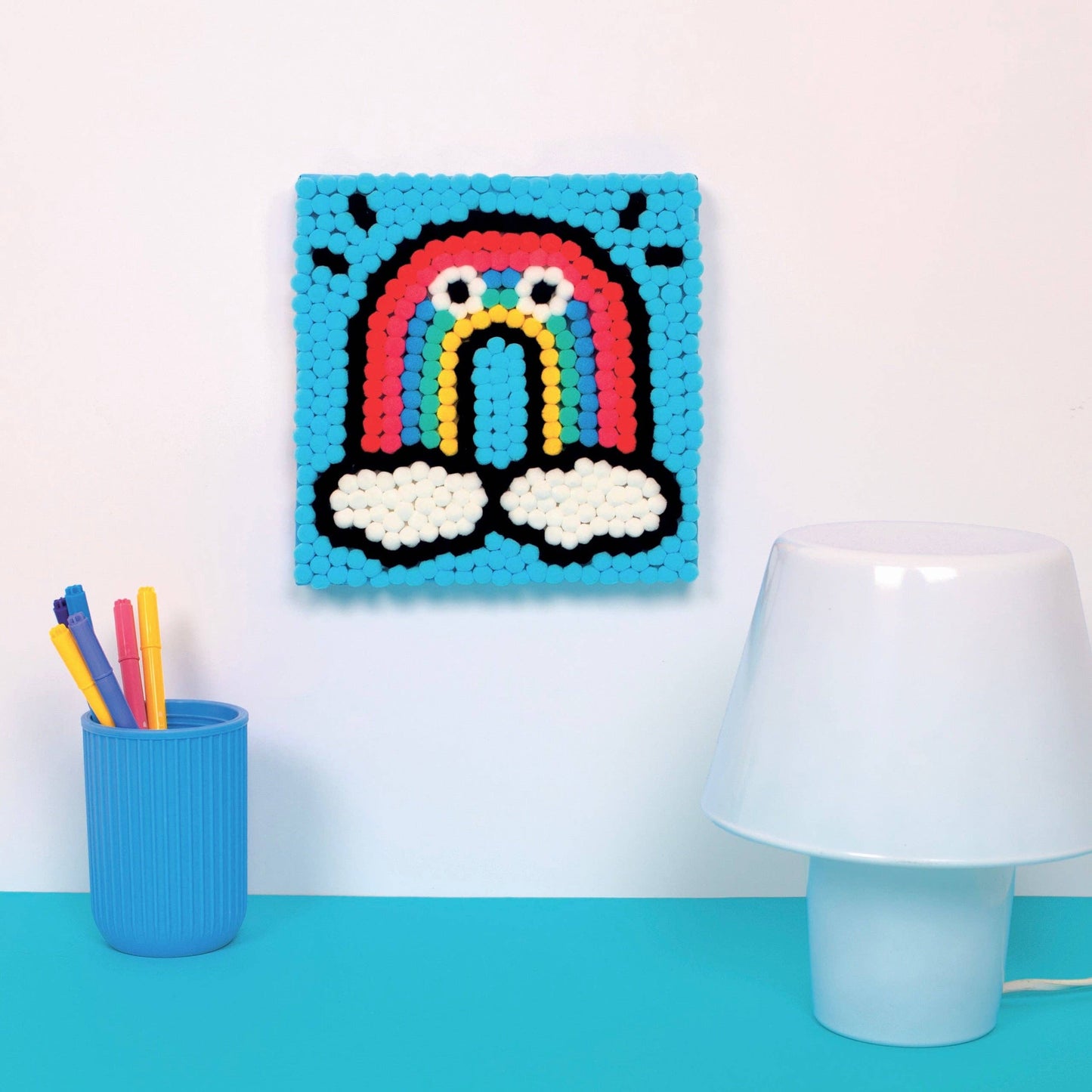 rainbow art piece on a wall with a lamp and cup of pens below.