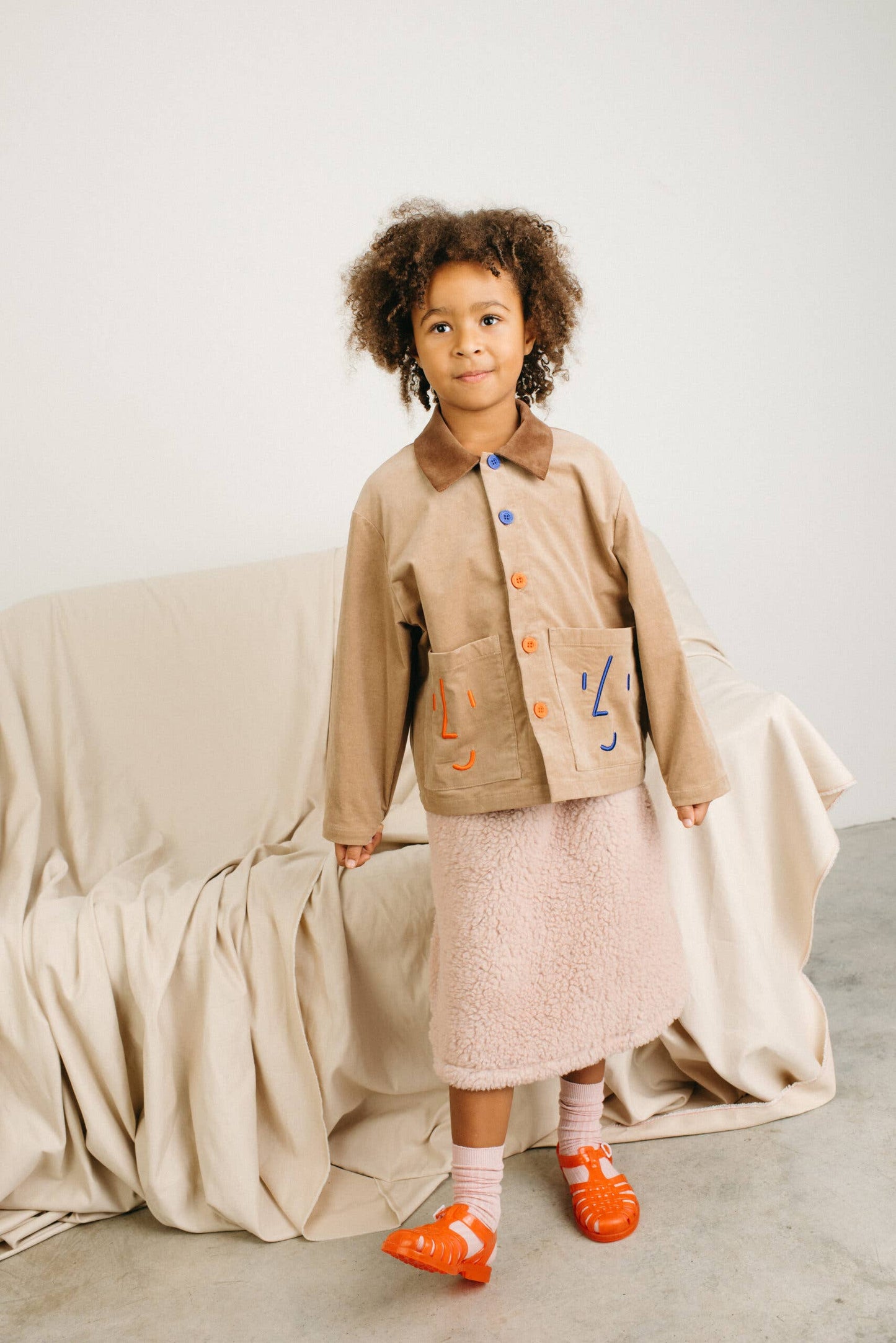 Child wearing a beige coat with colorful buttons, pink skirt, and orange shoes standing in a minimalistic room.