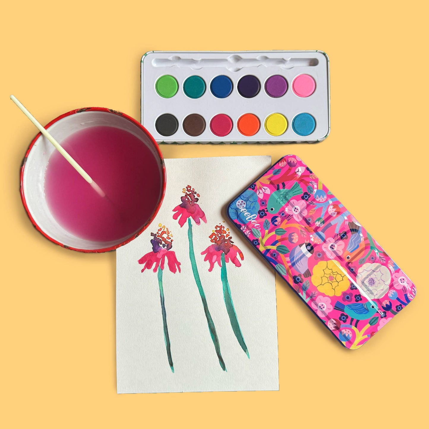 Colorful watercolor case with floral design next to watercolor painting and paint set on a yellow background