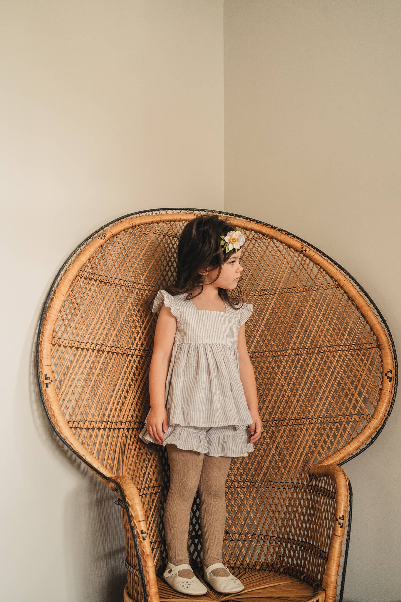 A toddler standing in a hanging rattan chair, wearing oat cable knit tights and a floral dress, with the chair taking up a large portion of the frame.
