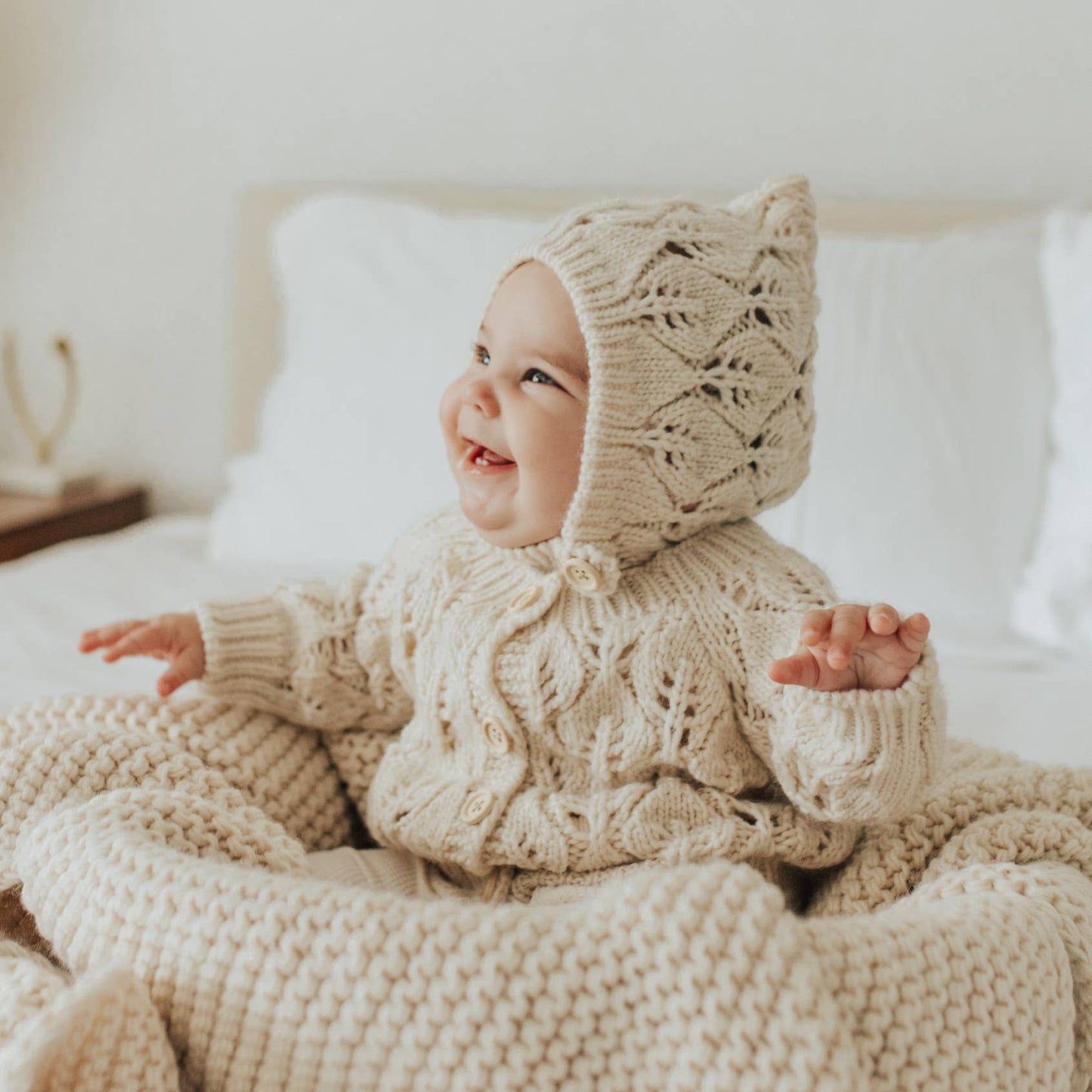 Baby wearing a beige knitted outfit sitting on a bed.