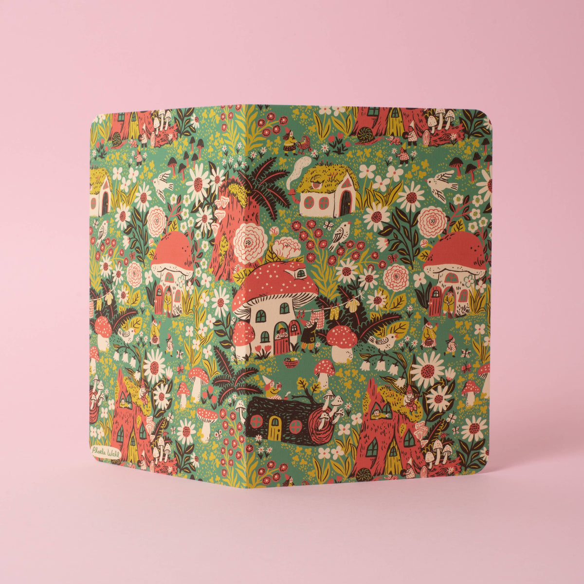 A softcover saddle-stitch notebook with vibrant wraparound cover featuring toadstool houses and flowers and gnomes, shown on a pink background