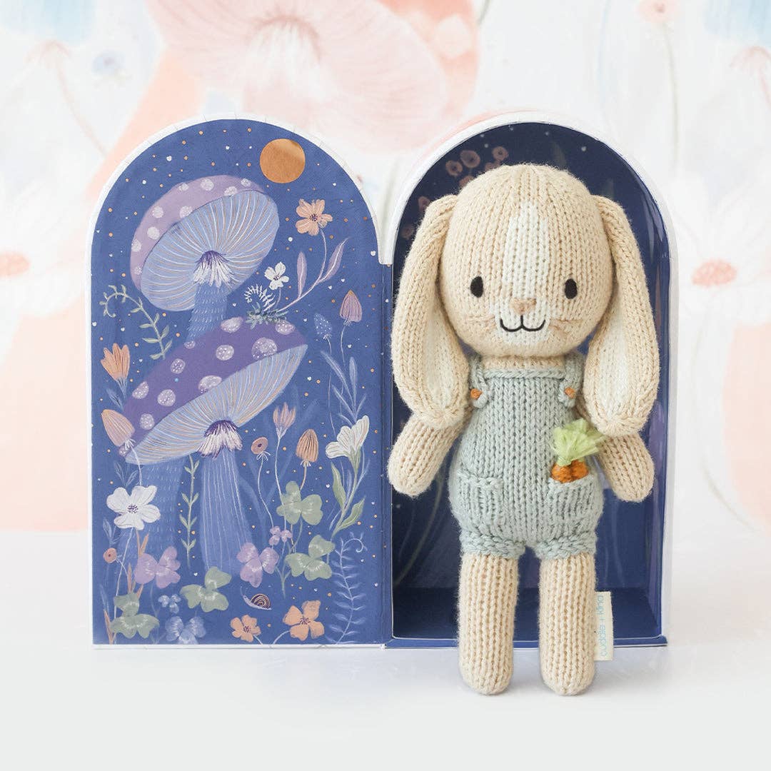 Knitted bunny toy in a blue outfit with a decorative box featuring floral and mushroom design.