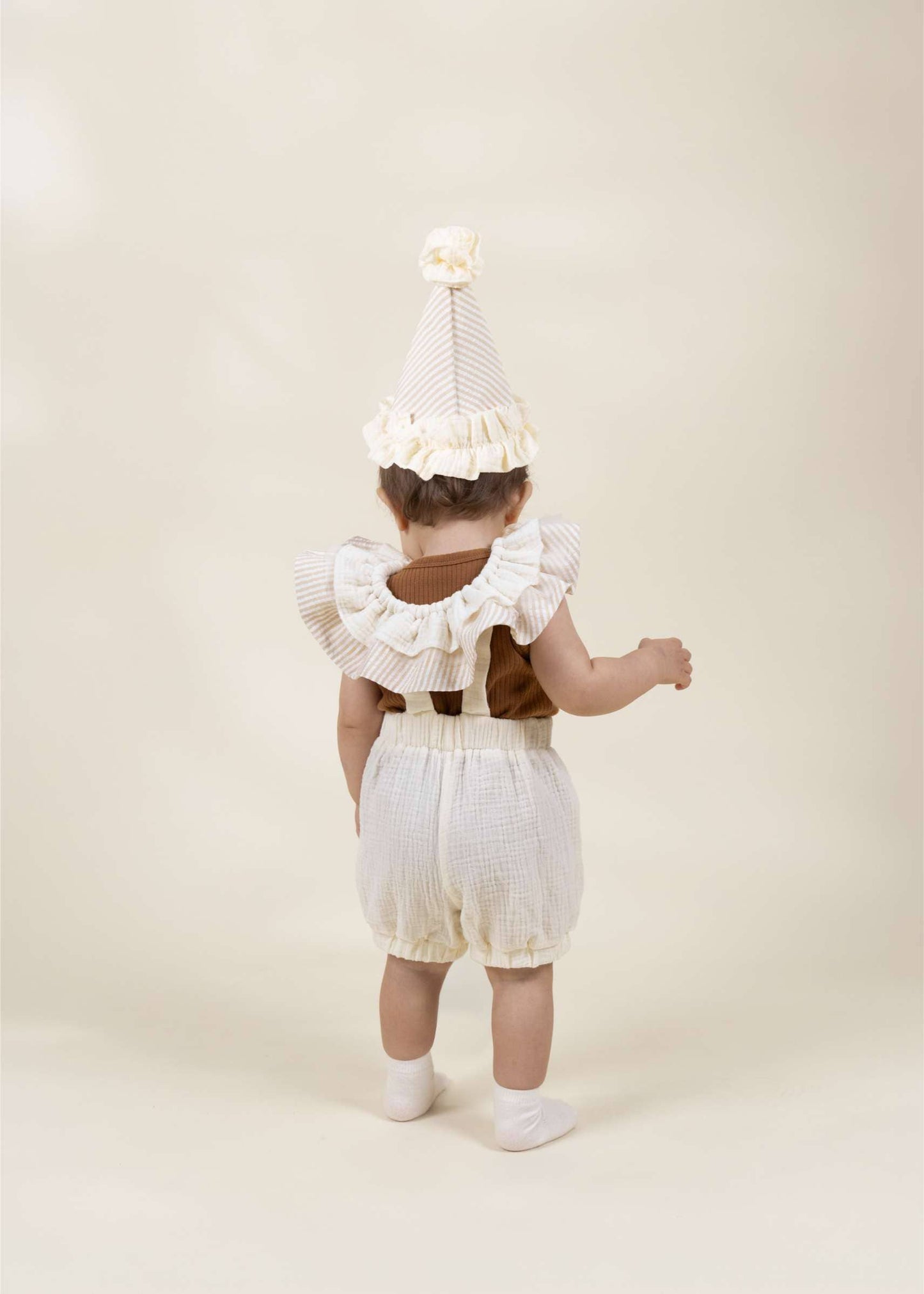 A baby viewed from behind wearing a cream and white striped party hat with a ruffled trim, a brown sleeveless top, and cream checkered bubble shorts, standing against a solid cream background.