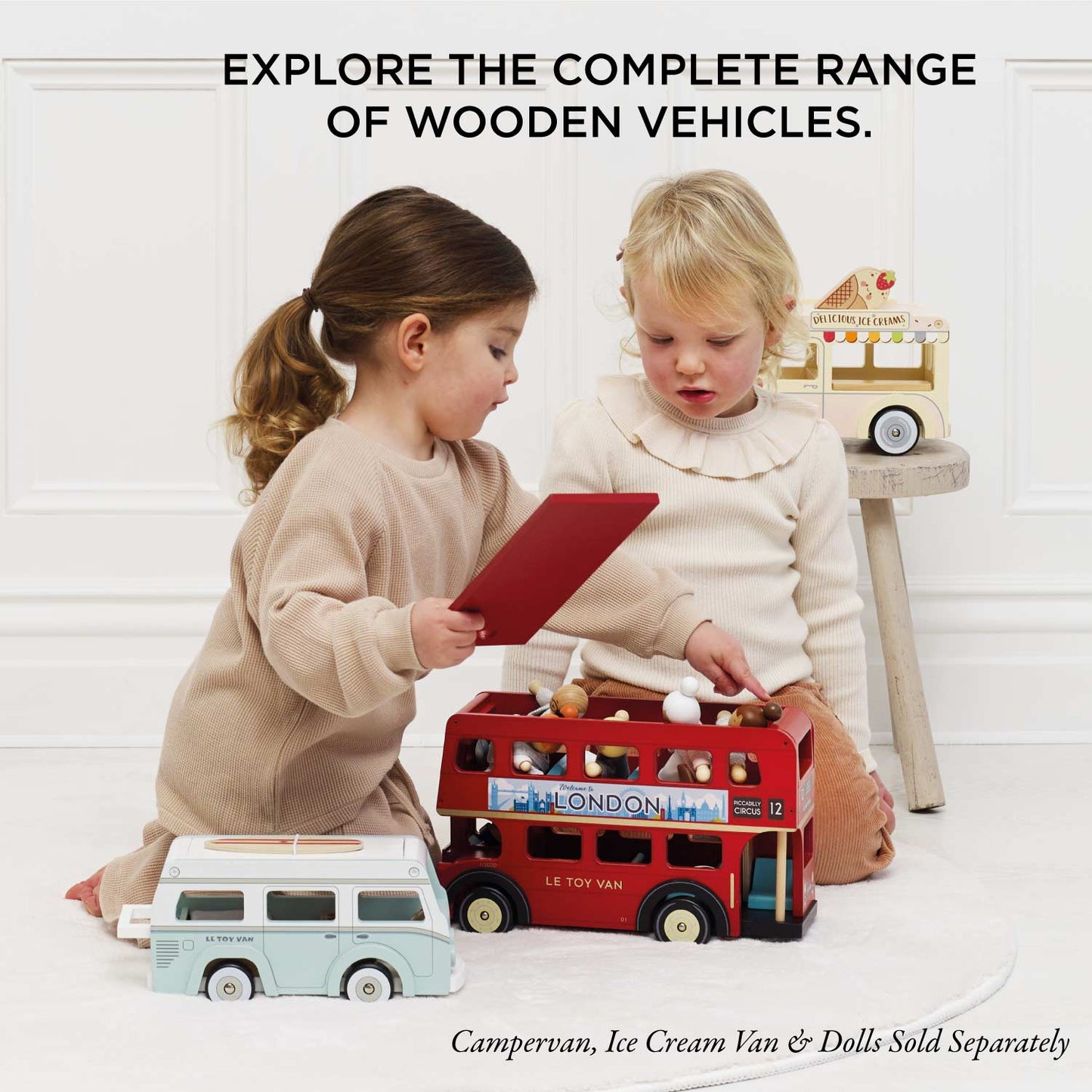 Two children playing with wooden toys including a London bus and campervan on a white floor.