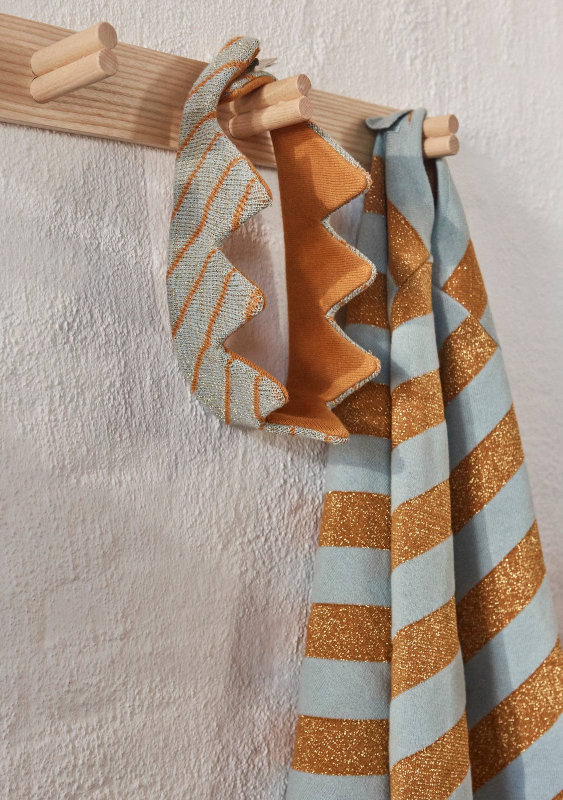 Textured fabrics of play crown and cape with geometric patterns hanging on a wooden rack against a textured wall.
