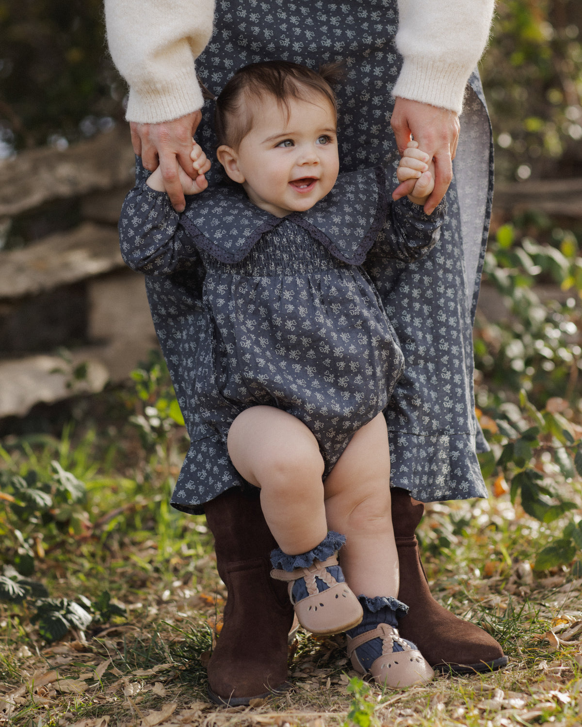 A baby wearing a dark blue and gray floral print romper, held up by an adult wearing brown boots and a cream sweater in an outdoor garden setting.