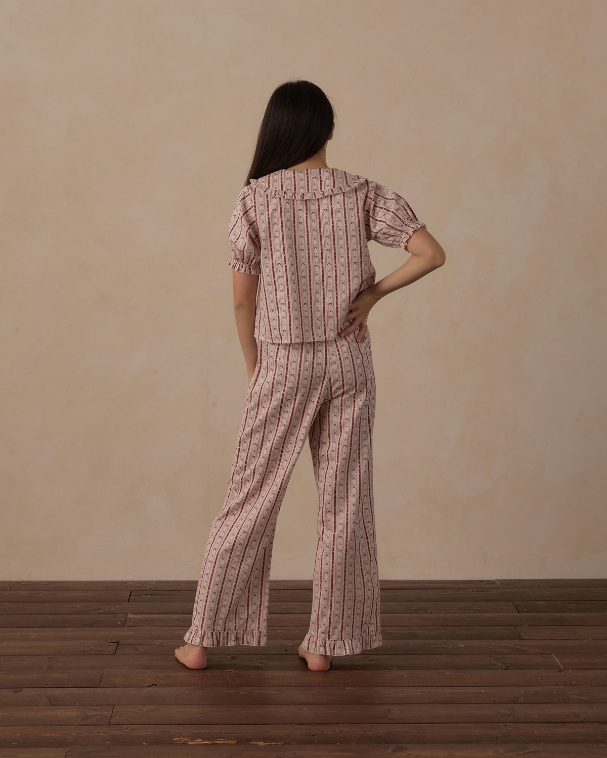 A woman stands with her back to the camera wearing a matching two-piece pajama set with short puff sleeves and wide-leg pants, featuring a pink and beige floral stripe pattern, set against a neutral beige wall and dark wood floor.