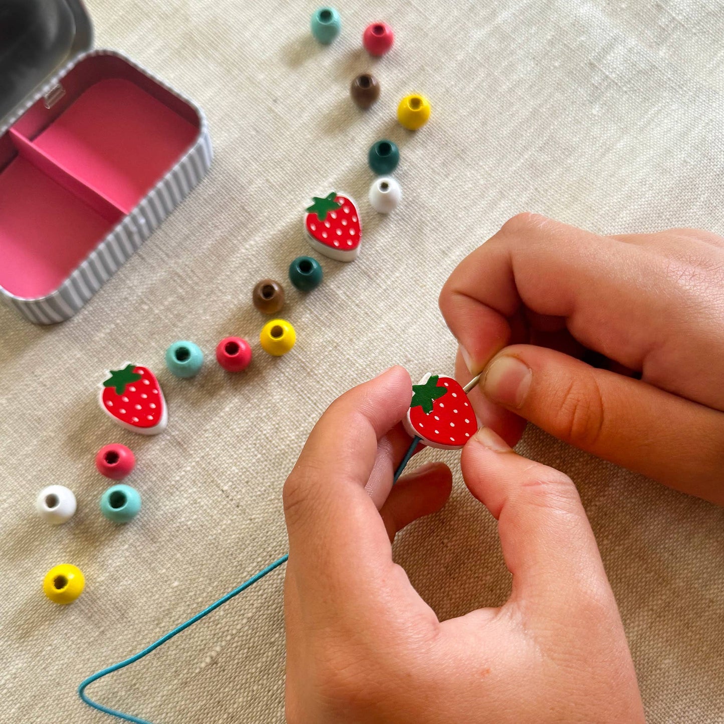 Child stringing colorful bead charms on a thread with a small pink container nearby.