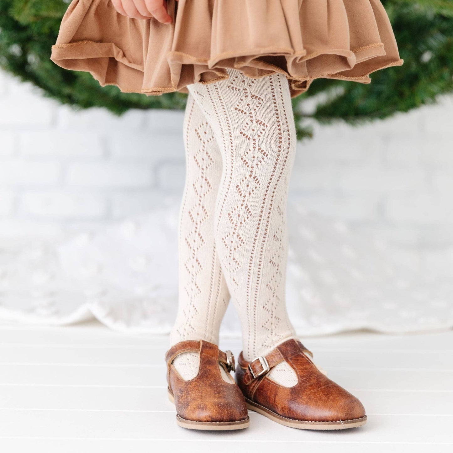 A toddler wearing brown shoes and vanilla knit tights with an open knit design, standing next to a decorative tree.