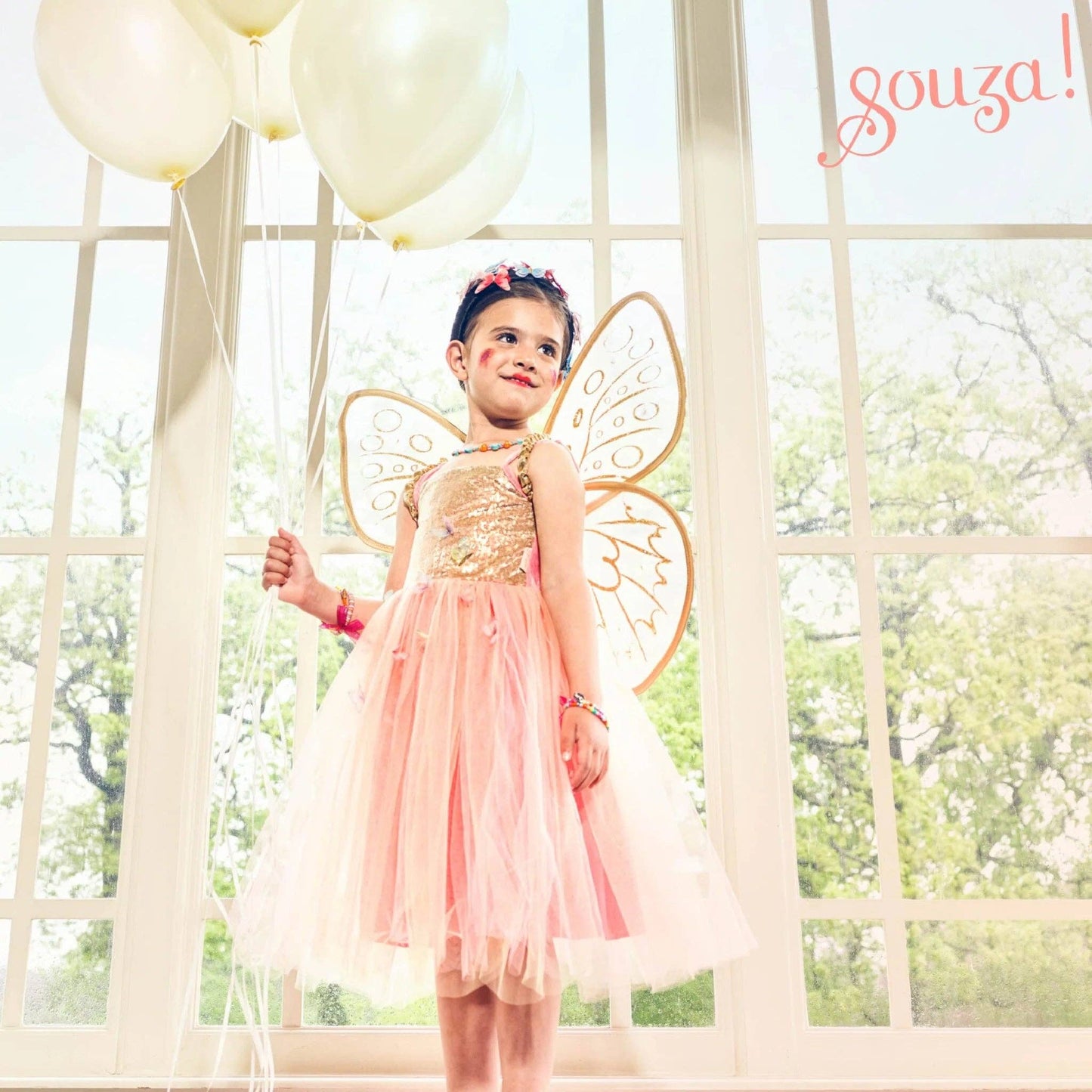 Young girl in a fairy costume with butterfly wings standing in front of a window with balloons.