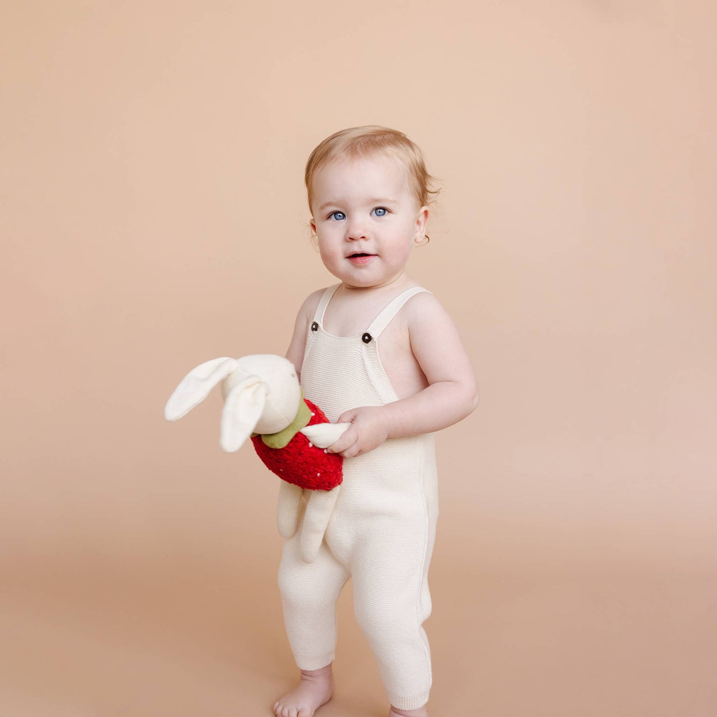 Child in white romper, holding a bunny toy, dressed as a strawberry