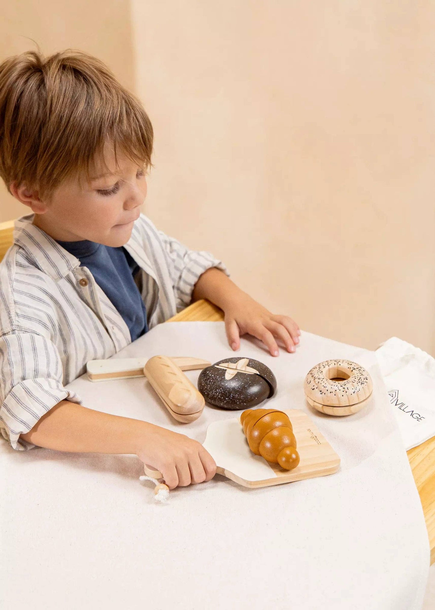 Child playing with a wooden bakery set including a variety of wooden breads and baking tools such as a knife and a cutting board, displayed in front of a canvas bag labeled 'COCO VILLAGE'.