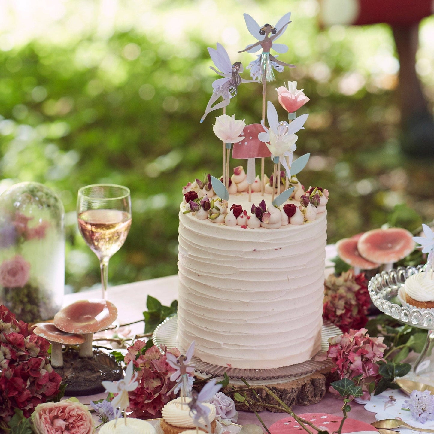 Decorative cake with fairy toppers on a table with flowers and a glass of wine.