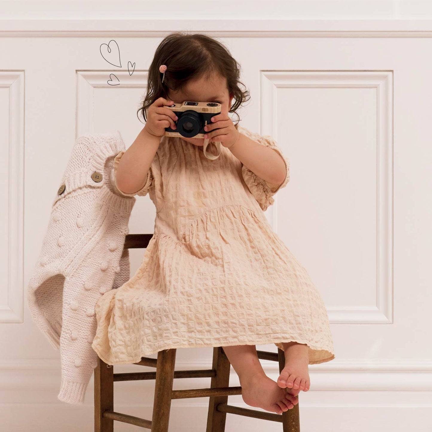 Child in a beige dress holding a camera against a white wall.