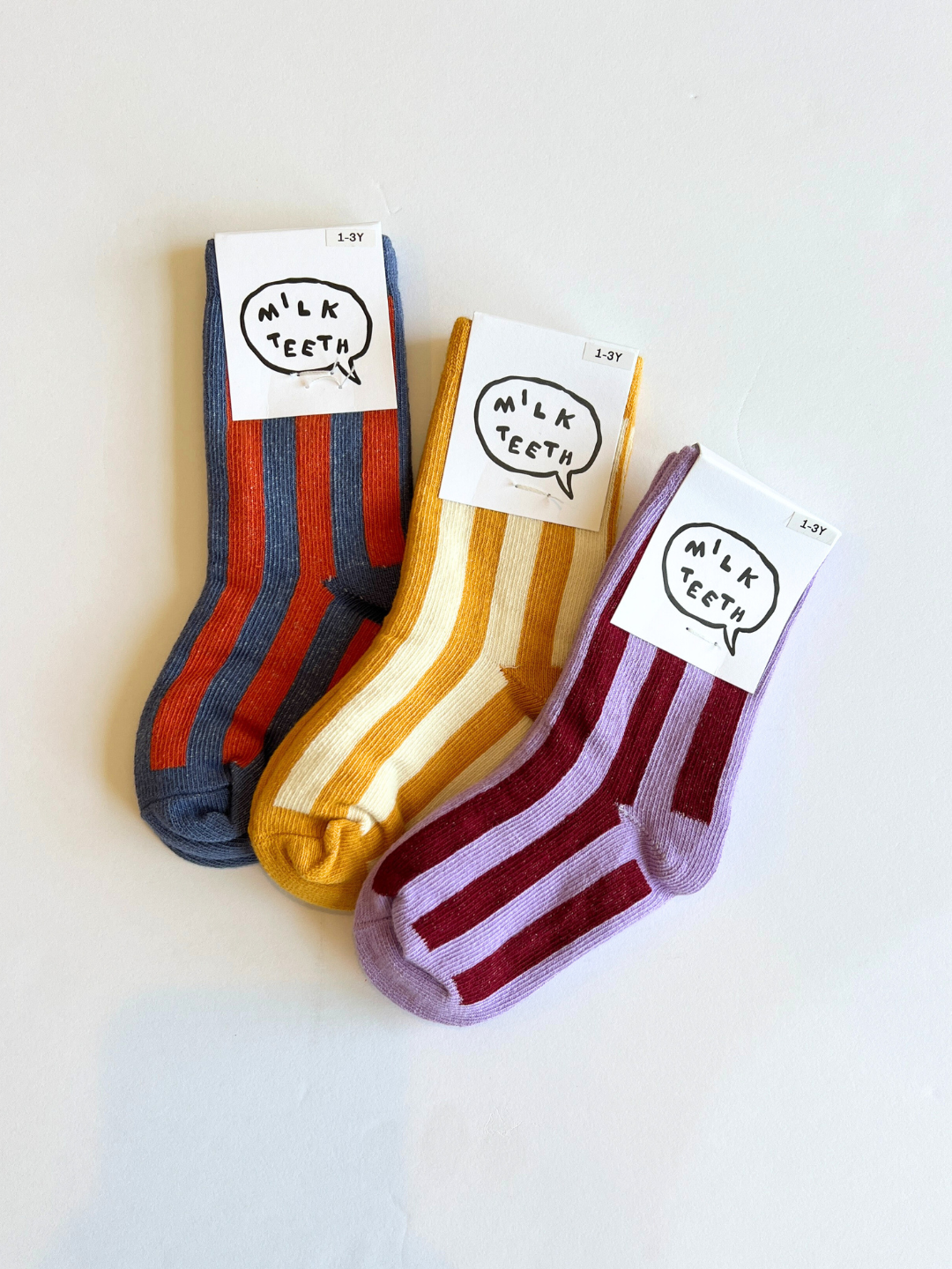 Three pairs of striped socks with 'Milk Teeth' labels on a light background