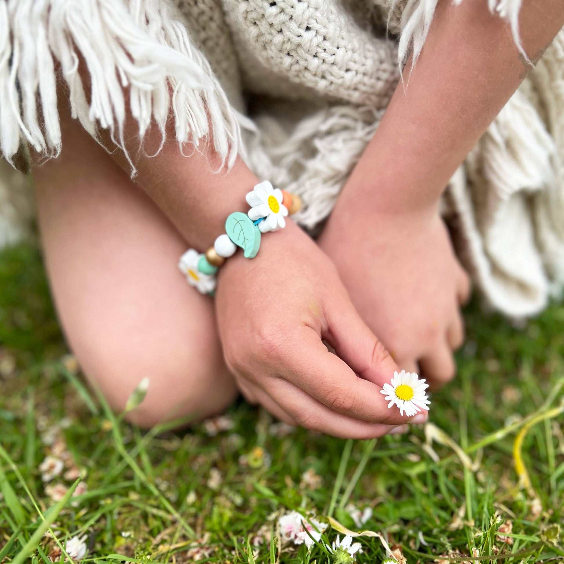 Child's hands, holding a daisy, wearing a beaded bracelet with flowers on a grassy background