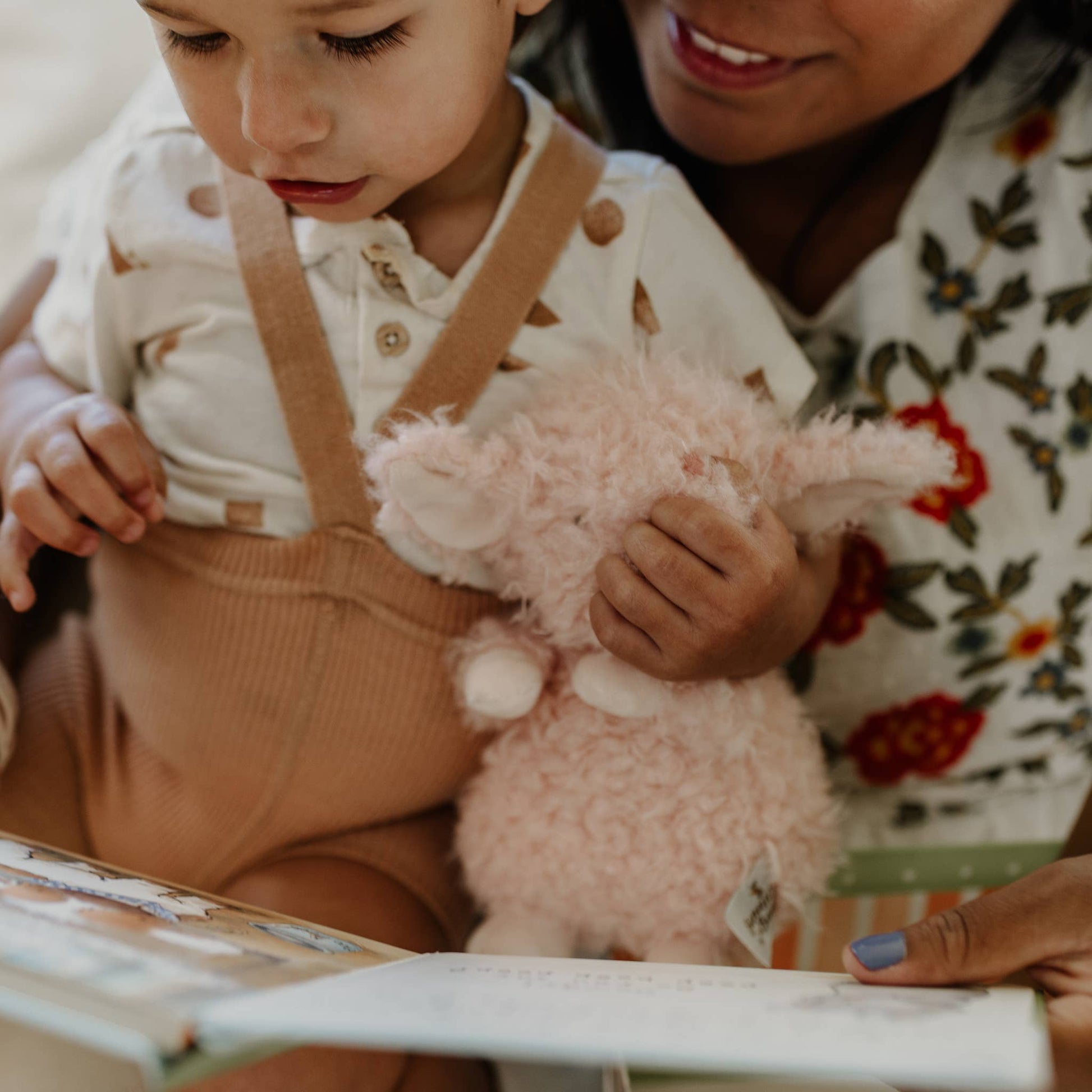 Child holding a pink fluffy toy with an adult next to them, both looking at a book.