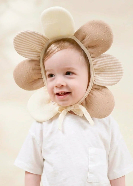 A smiling baby wearing a soft beige flower-shaped bonnet with ribbed petals and a cream bow, paired with a white short-sleeve shirt against a warm cream background.
