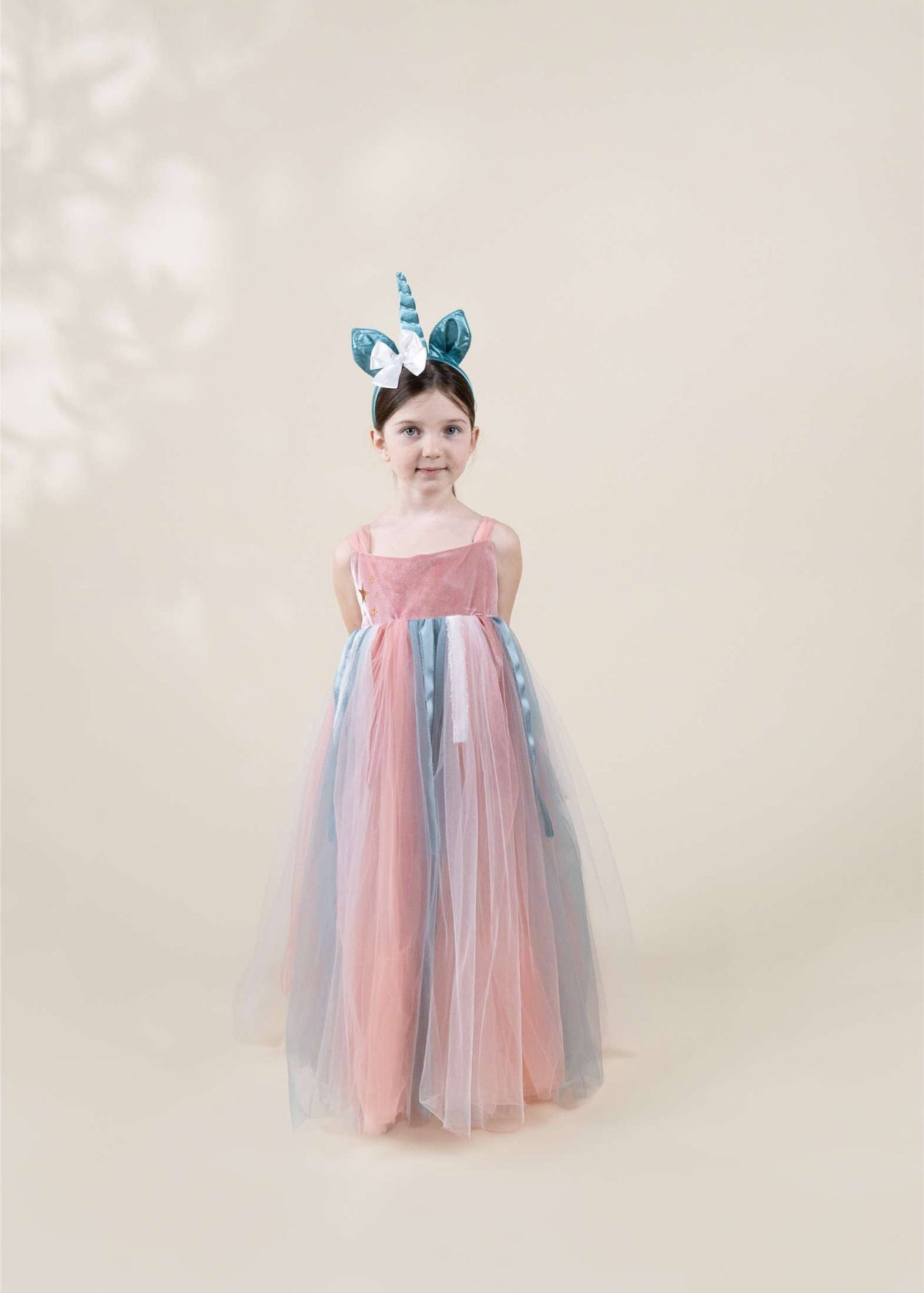 A young girl stands against a soft beige studio background wearing a long tulle dress with pink, blue, and gray panels, plus a matching horn headband.