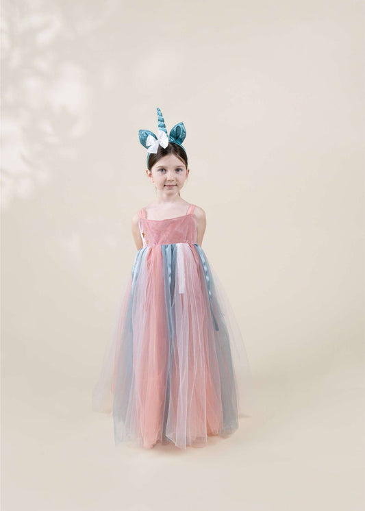 A young girl stands against a soft beige studio background wearing a long tulle dress with pink, blue, and gray panels, plus a matching horn headband.