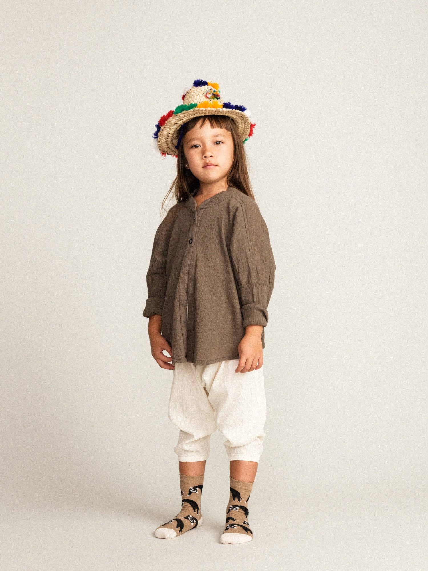 Child wearing a brown shirt, white pants, and a colorful hat and brown cat socks on a plain background