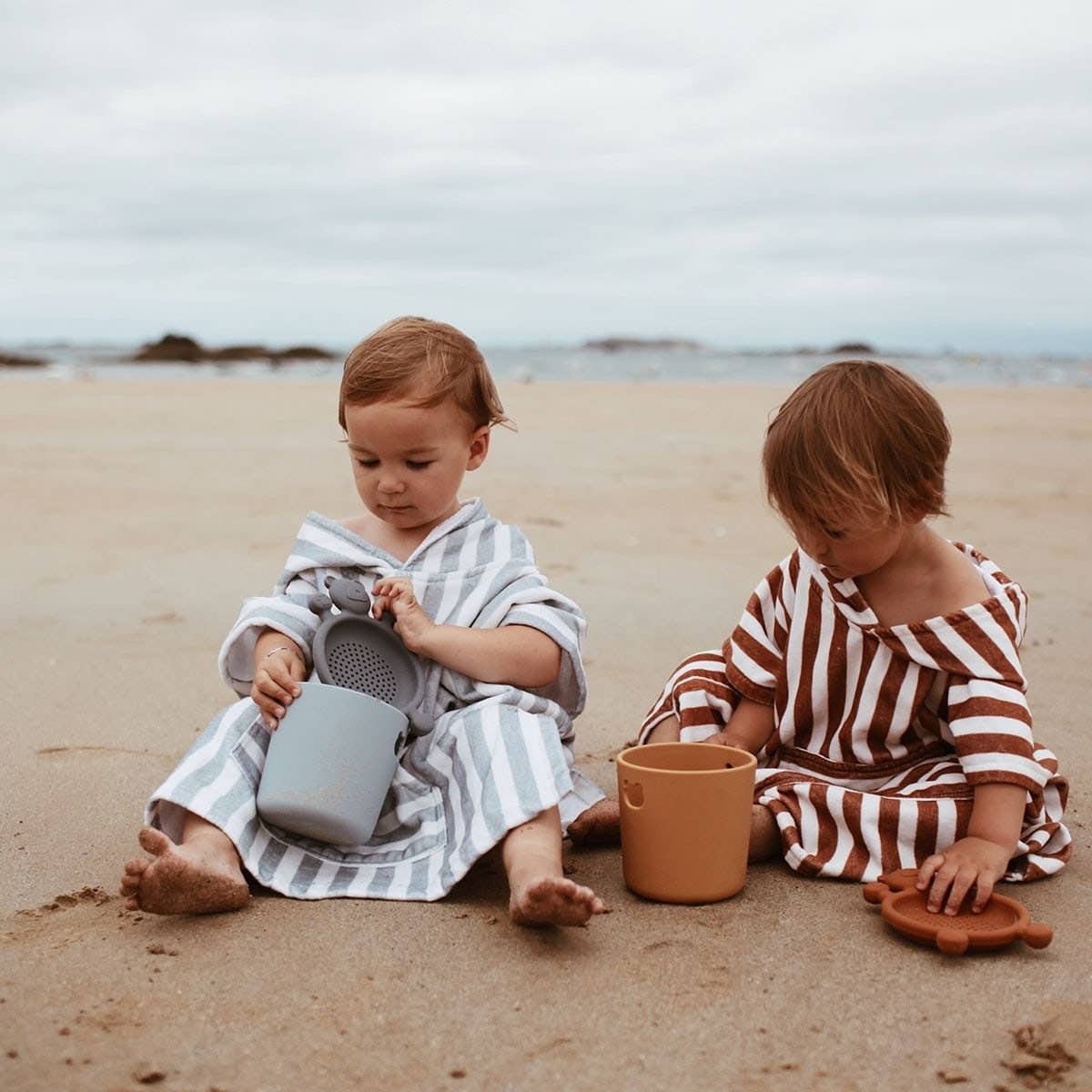 Two children playing on a beach with sand toys, wearing hooded, striped towels.
