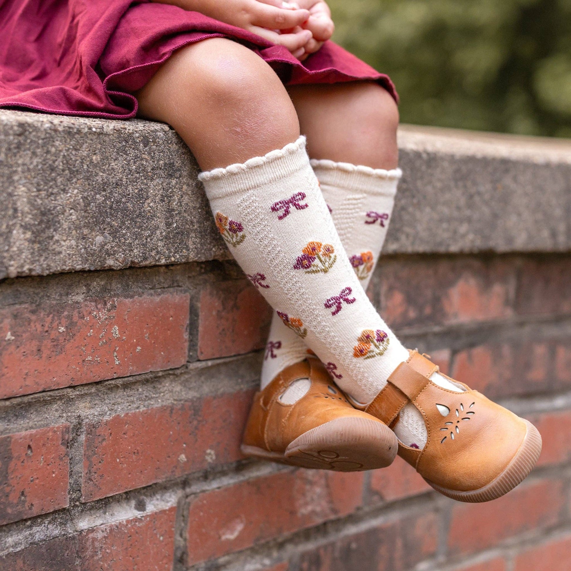 Child wearing white socks with floral patterns and brown shoes sitting on a brick wall.