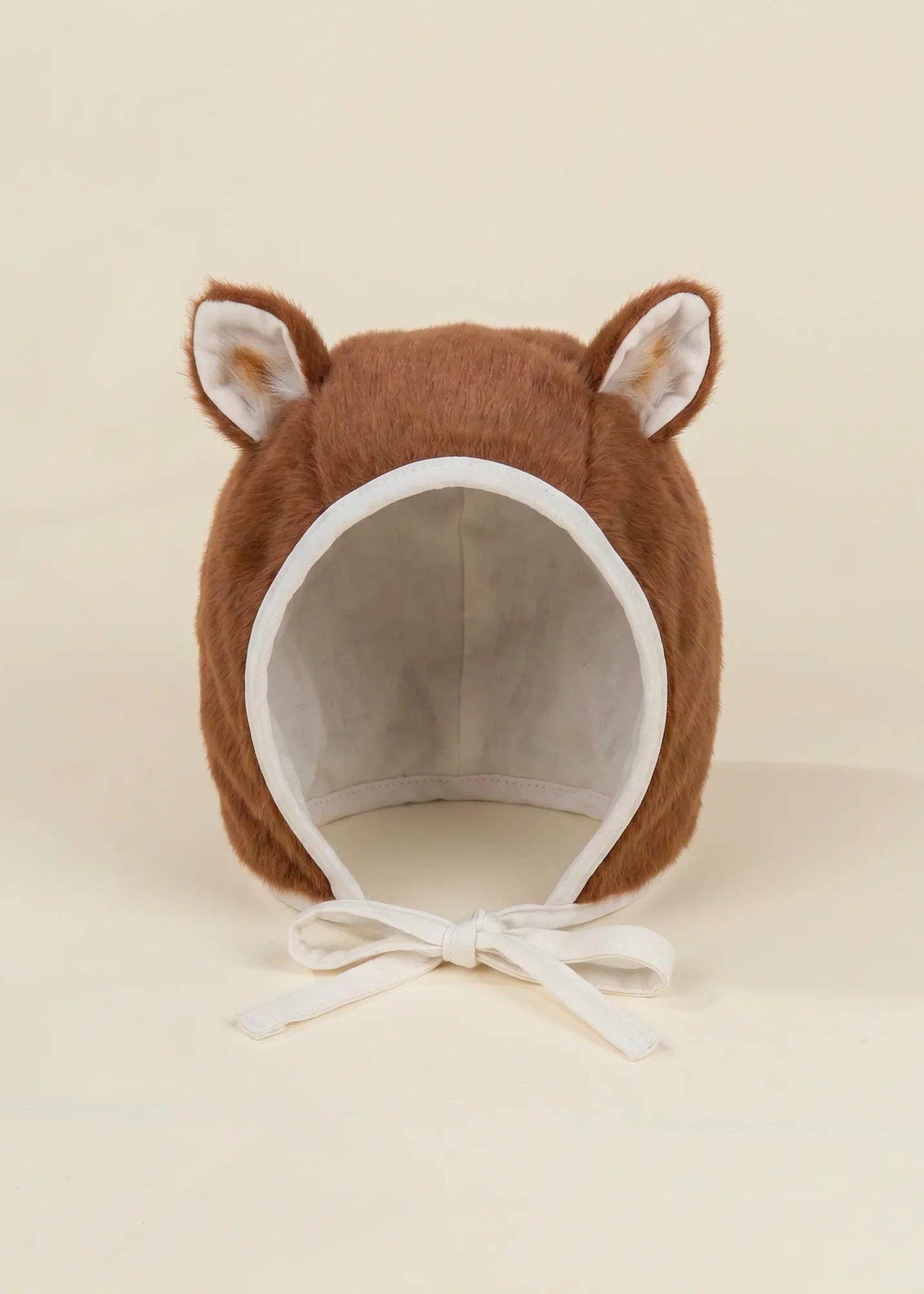 A brown plush baby bonnet with upright fox ears and white chin ties, photographed against a plain warm cream background.