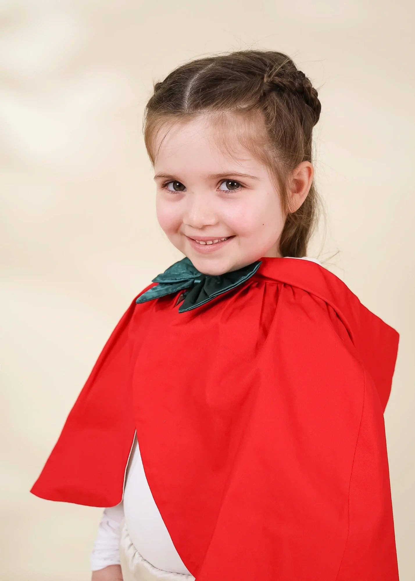 A young girl with brown hair in a half-up braid, wearing a bright red cape with a dark green bow accent, posing against a soft beige studio background.