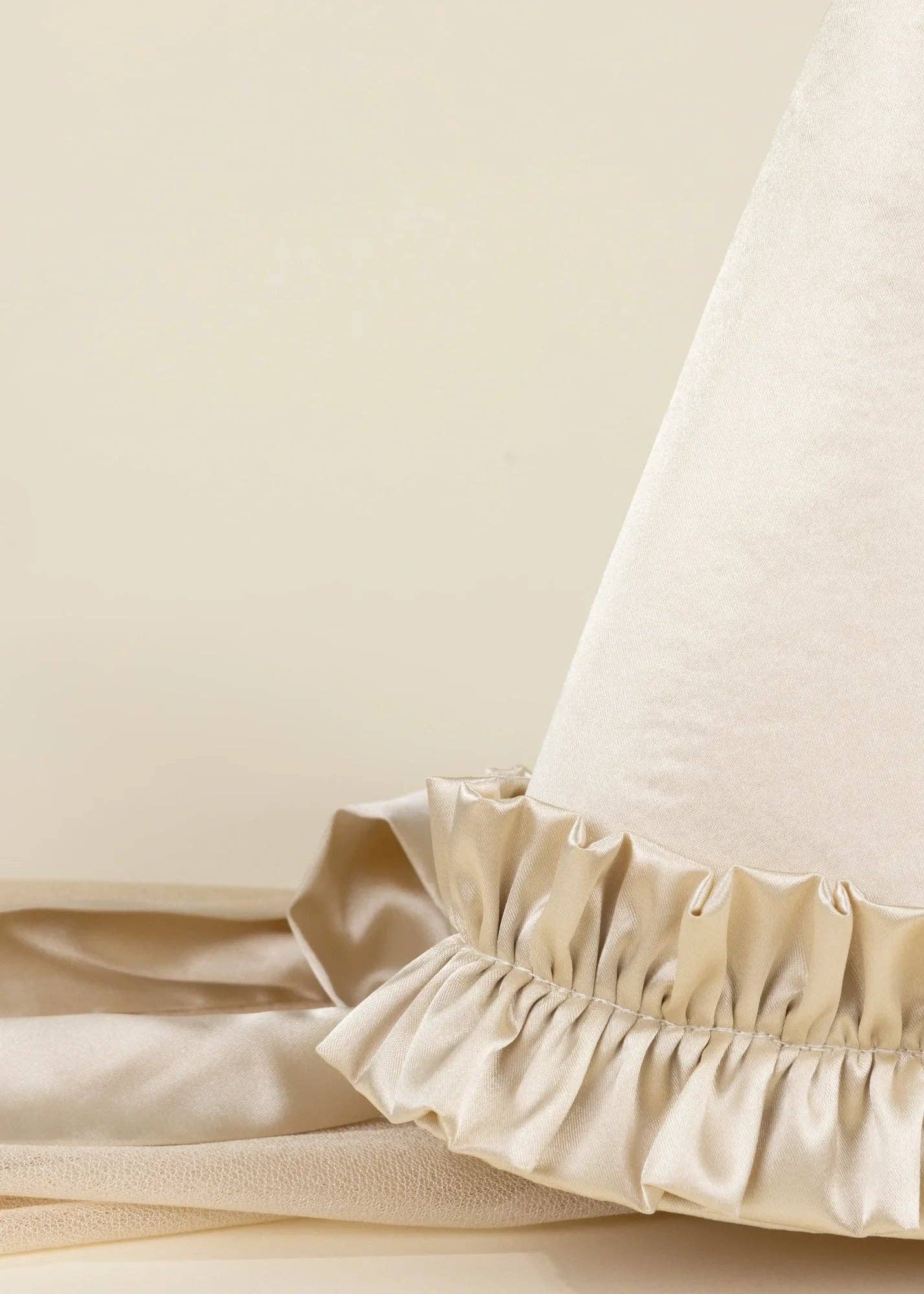 Close-up of a cream-colored hat-like textile with a gathered ruffled trim, resting on a matching satin fabric against a warm beige background.