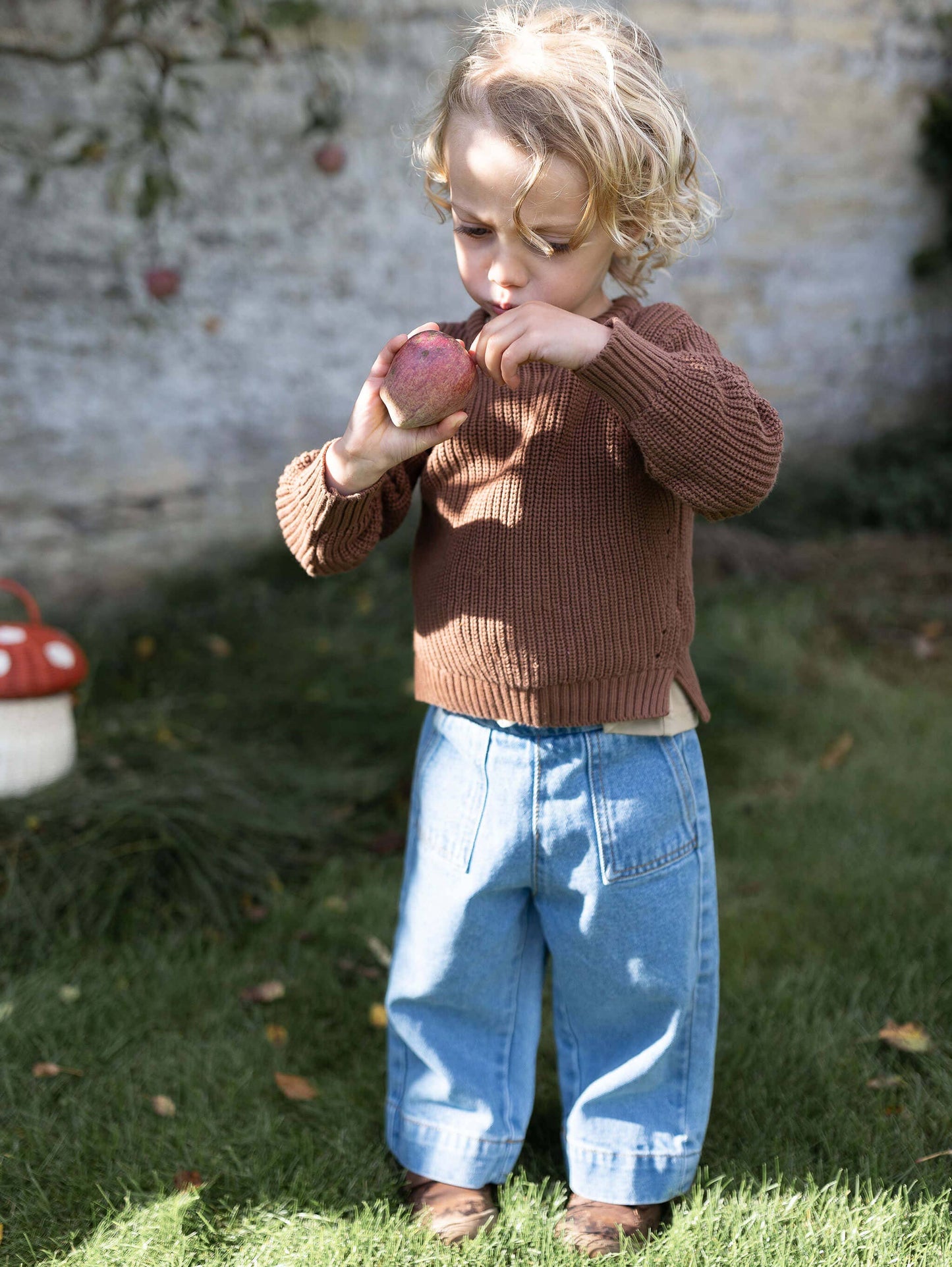Child in a brown sweater and blue jeans eating an apple outdoors.