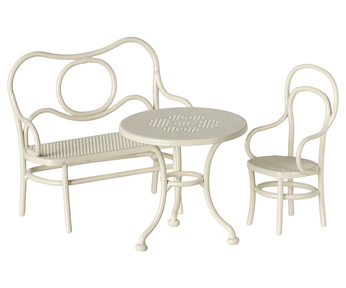 A dollhouse sized three-piece outdoor patio bistro set in a warm off-white finish, featuring a curved metal bench with a slatted seat, a matching armchair, and a small round table with a slatted top.