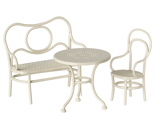 A dollhouse sized three-piece outdoor patio bistro set in a warm off-white finish, featuring a curved metal bench with a slatted seat, a matching armchair, and a small round table with a slatted top.