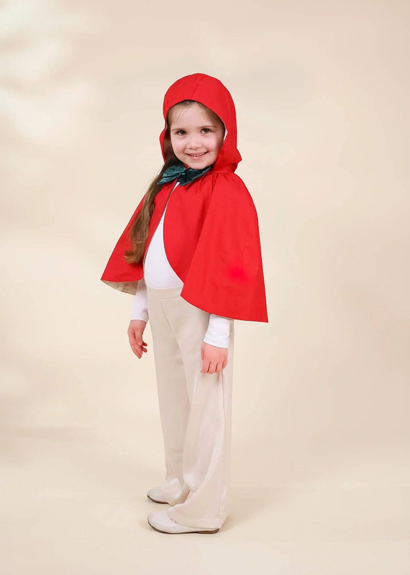A young girl modeling a red hooded cape costume over a white outfit, standing against a plain beige studio background.