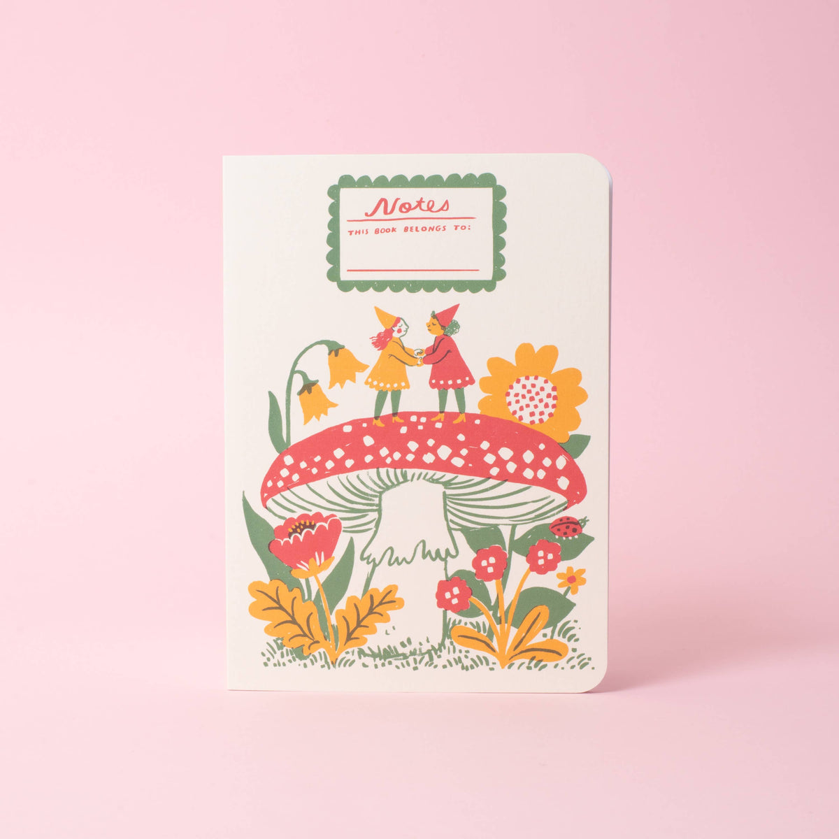 A softcover saddle-stitch notebook with vibrant wraparound cover featuring two gnomes on top of a red toadstool, on a pink background