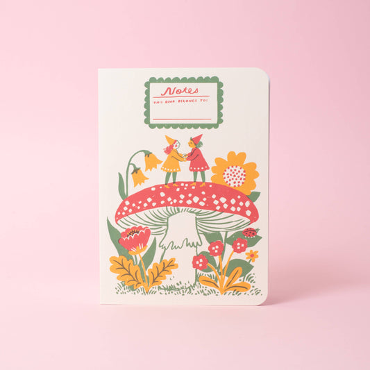 A softcover saddle-stitch notebook with vibrant wraparound cover featuring two gnomes on top of a red toadstool, on a pink background