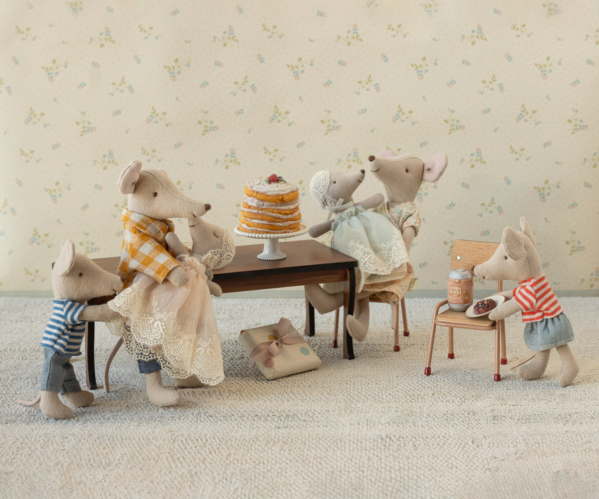 Stuffed animal family gathered around a small table with a cake, set against a floral wallpaper background.