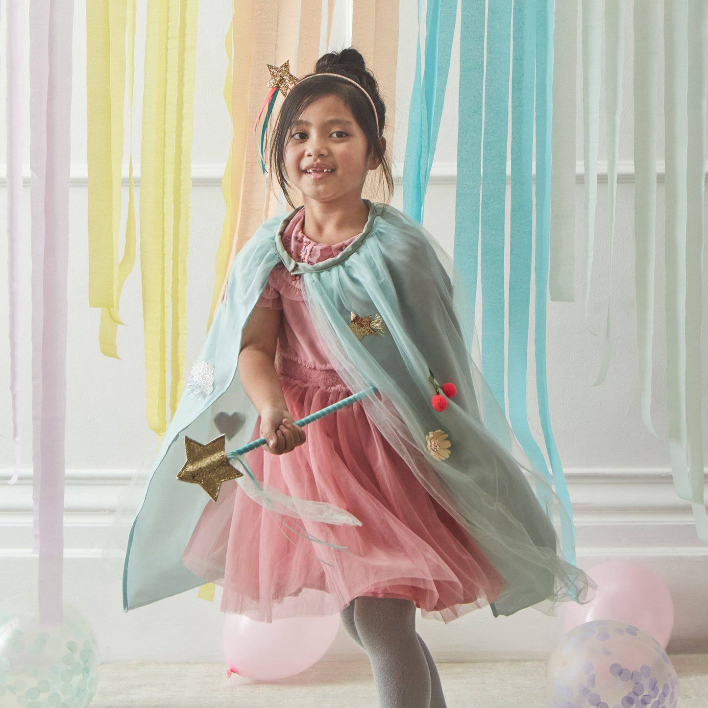 Young girl in a pink dress with a light green cape and wand, standing in front of colorful curtains.