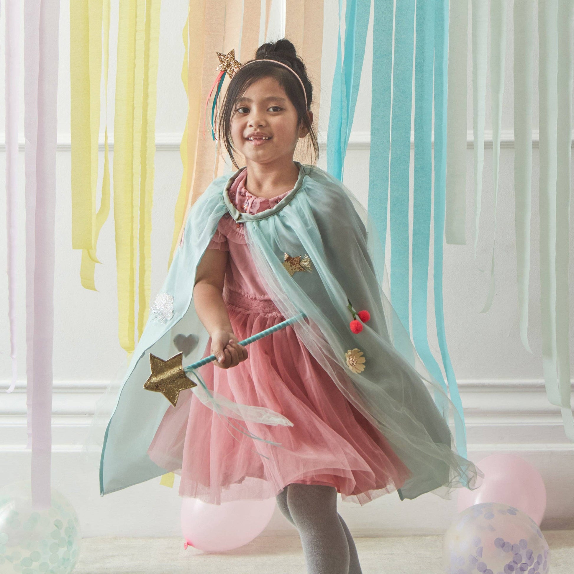 Young girl in a pink dress with a light green cape and wand, standing in front of colorful curtains.