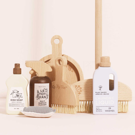 Set of wooden play cleaning products including bottles, a scrub brush, and a dustpan on a light background