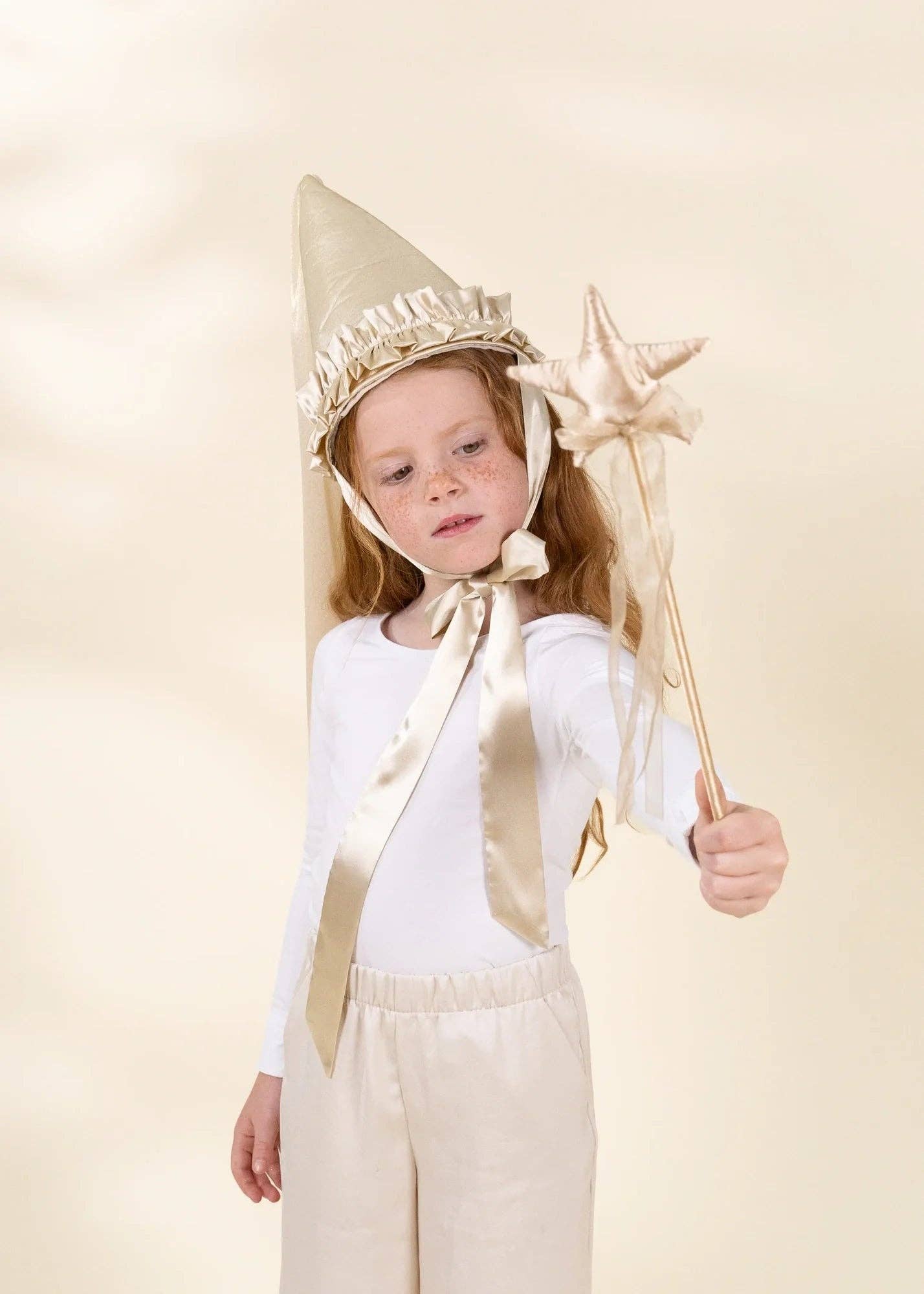 A young girl wearing a gold and white fairy costume, featuring a pointed gold hat with a white ruffled trim, a matching satin ribbon, and holding a gold star wand, set against a plain beige studio background.