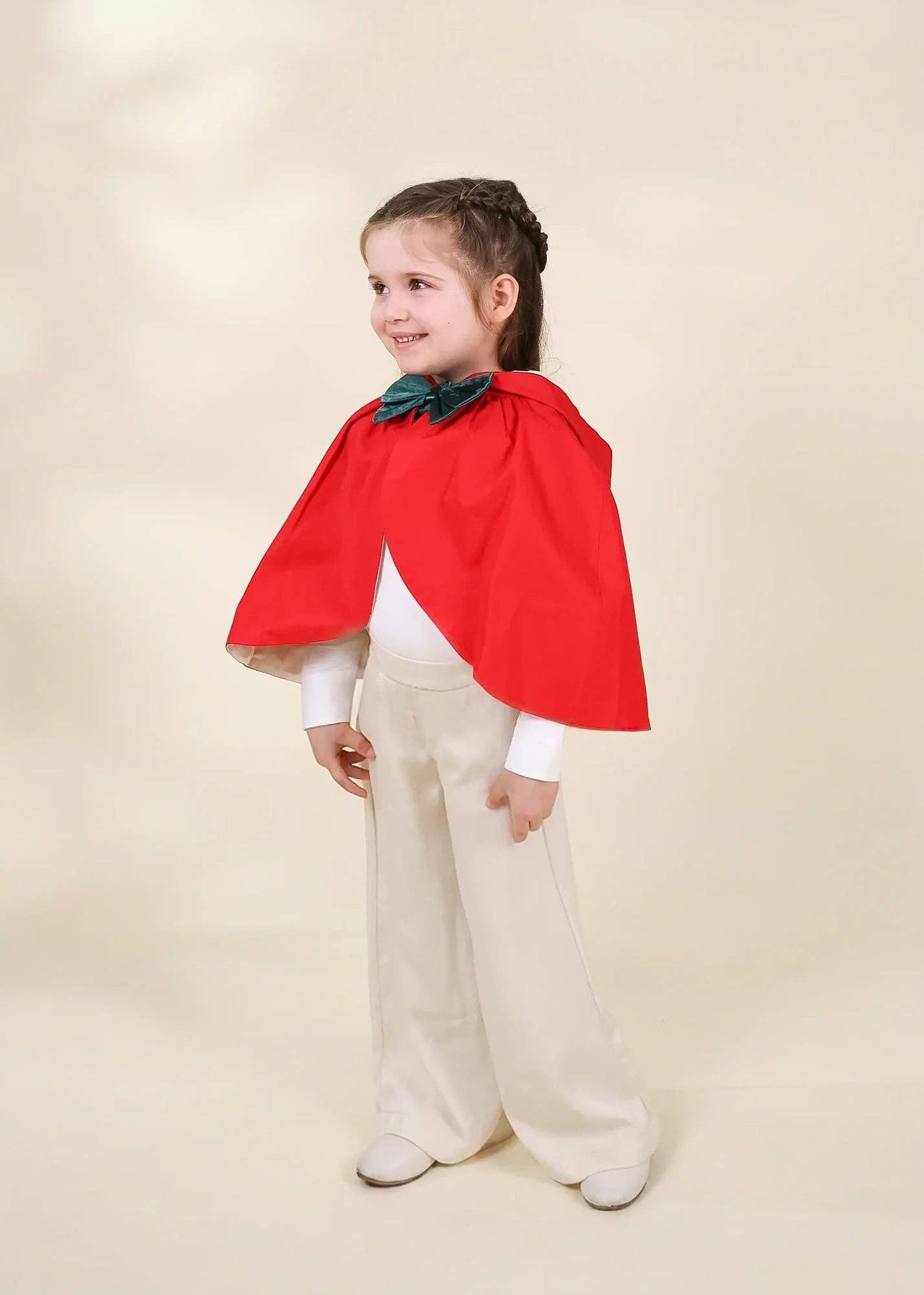 A young girl modeling a children's costume consisting of a bright red cape, a dark green bow tie, a white long-sleeve shirt, and cream-colored trousers, set against a solid beige studio background.