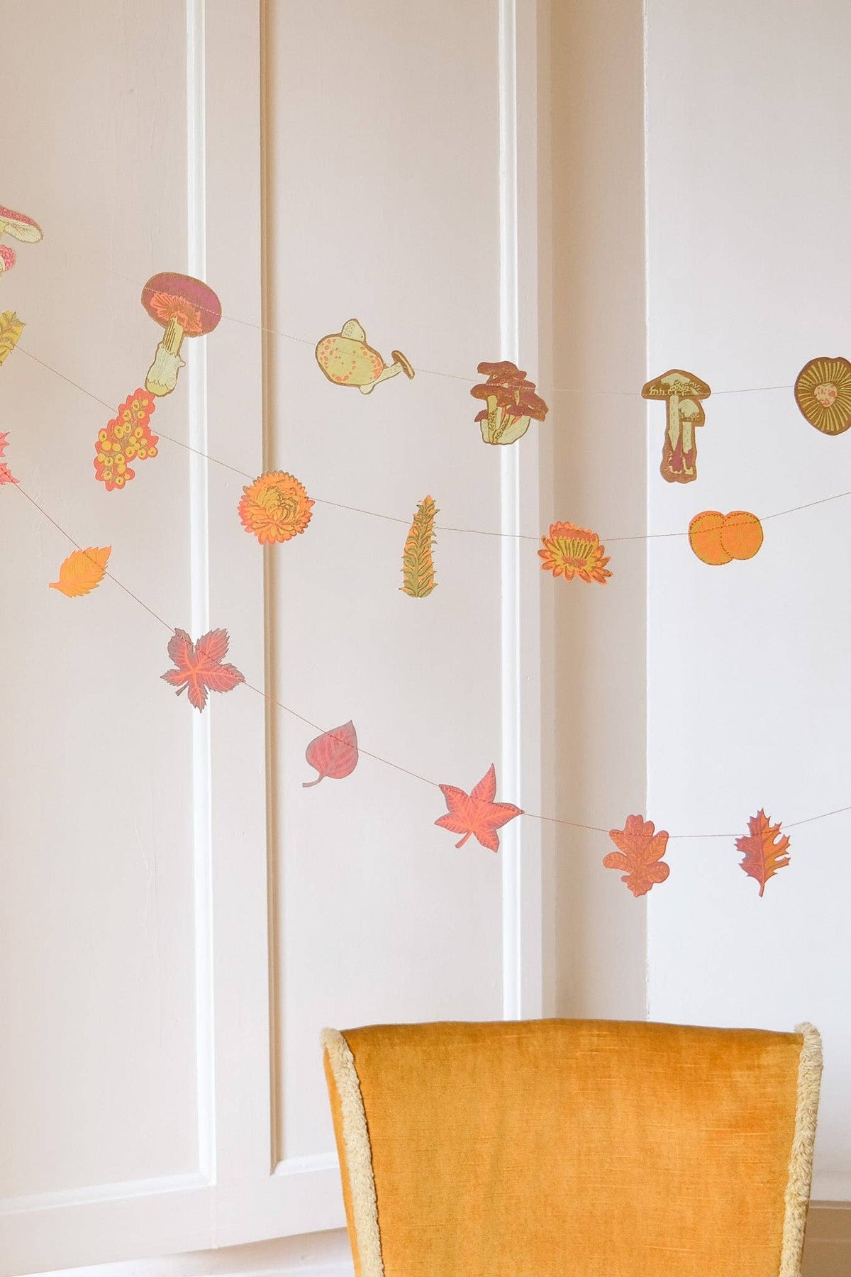 Mushroom, pumpkin and autumn leaf paper garlands hanging across a paneled wall above a yellow chair.