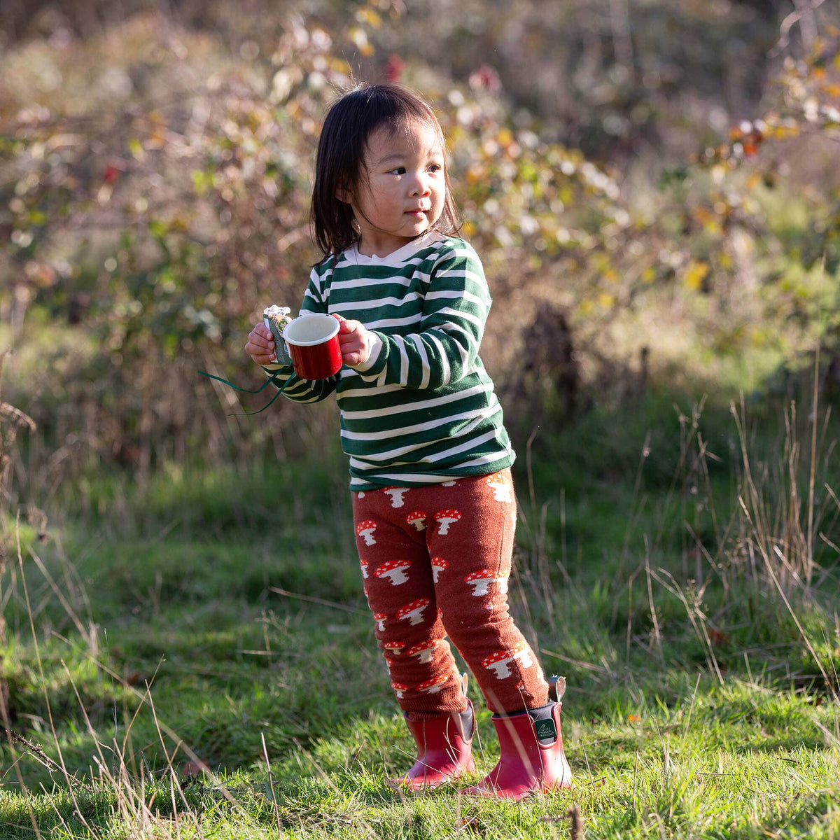 Child wearing a green and cream striped long-sleeve T-shirt outdoors with mushroom joggers.
