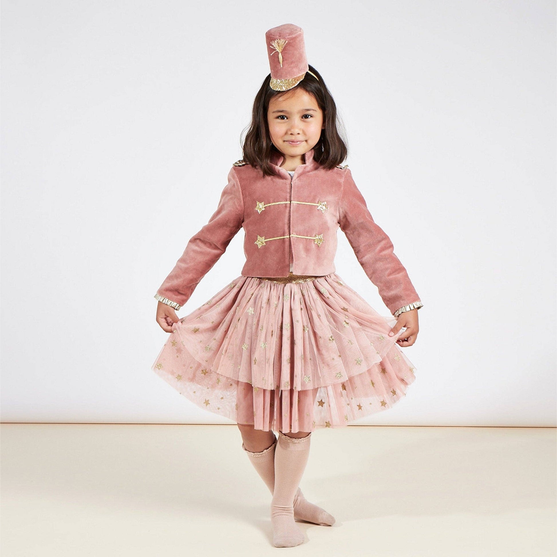 A young girl modeling a dusty pink ballet tutu costume set, which includes a velvet jacket with gold buttons, a matching tiered tulle skirt with gold embroidery, a pink tutu hat, and pink tights, set against a plain light background.