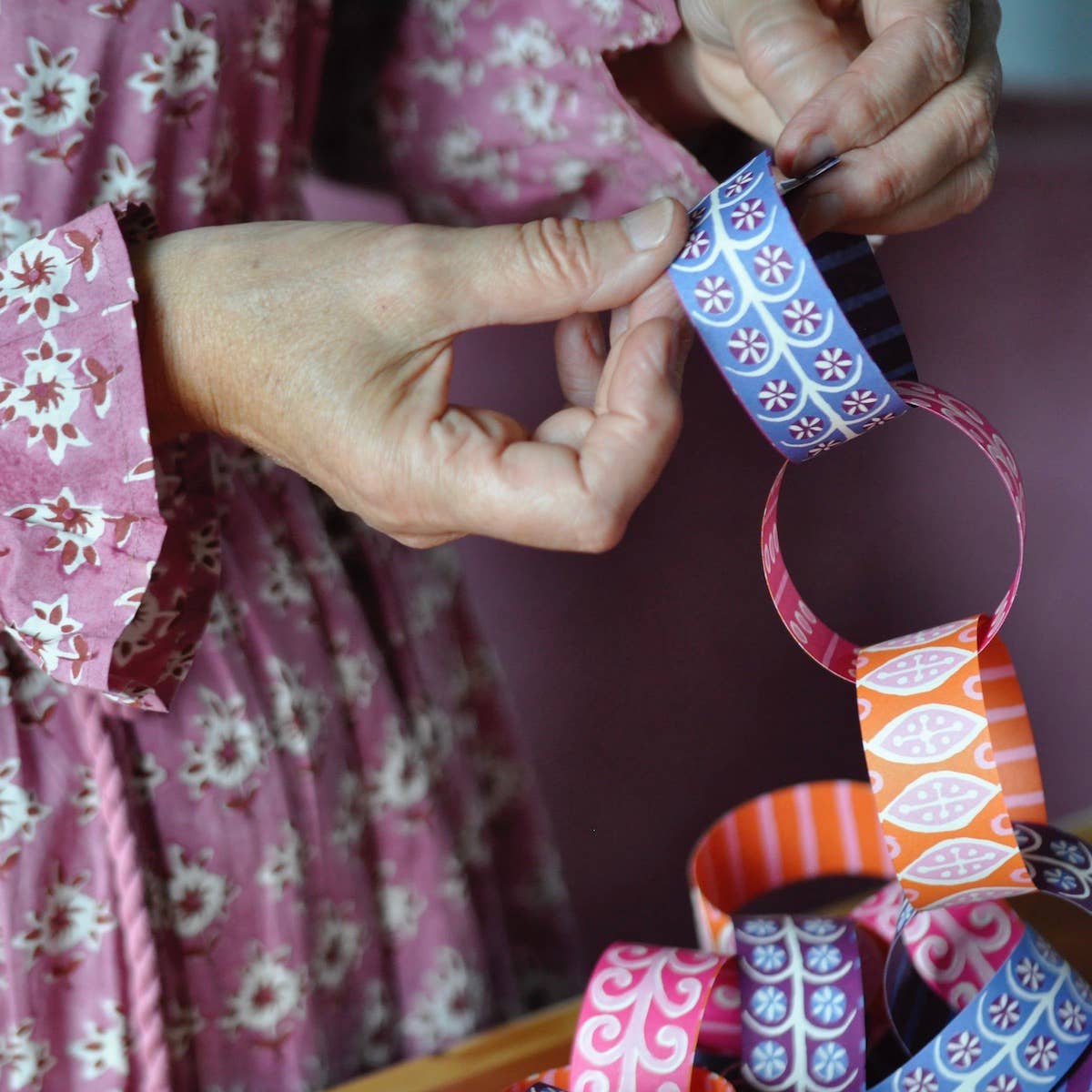 Hands joining a blue patterned paper link to a chain of pink and orange links.