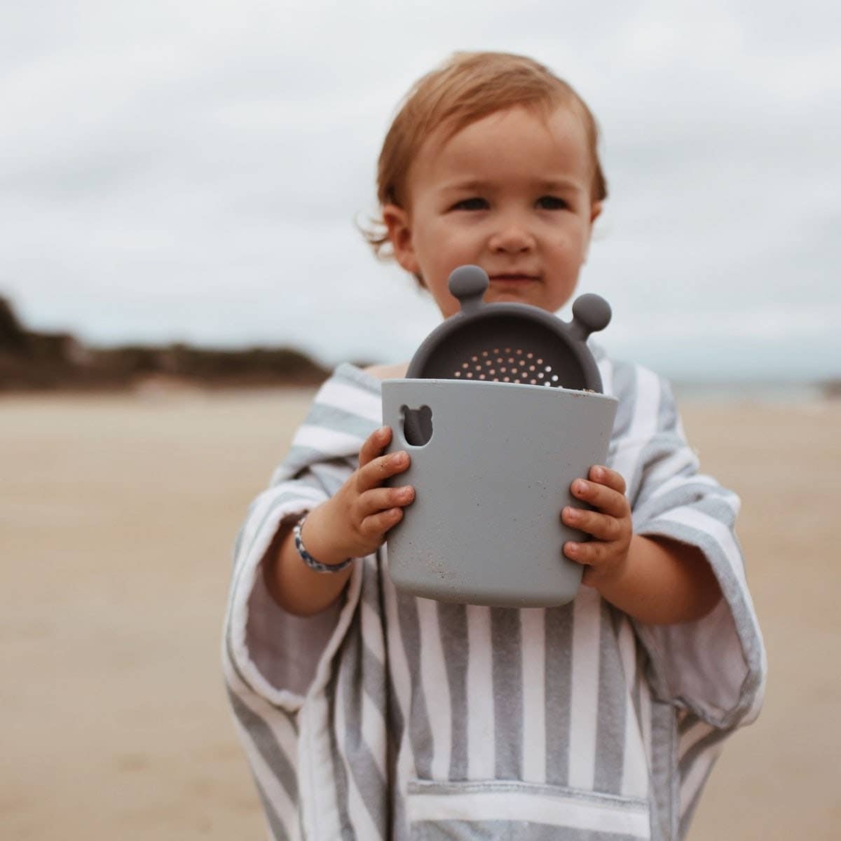 Child holding a gray sand toy with a textured striped towel on a beach