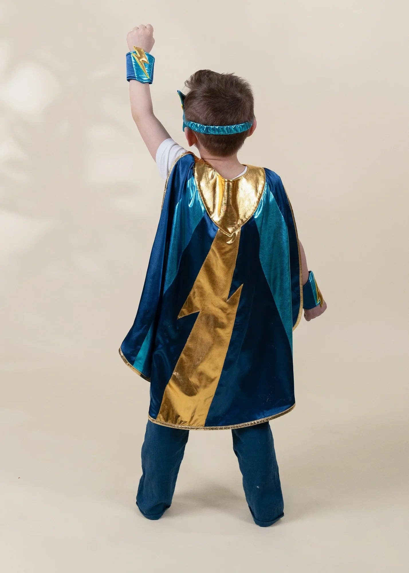 A child wearing a superhero cape costume, shown from the back. The costume features a shiny gold lightning-bolt design on a dark blue base, with matching blue pants and accessories including a headband and wristbands, set against a plain beige studio background.